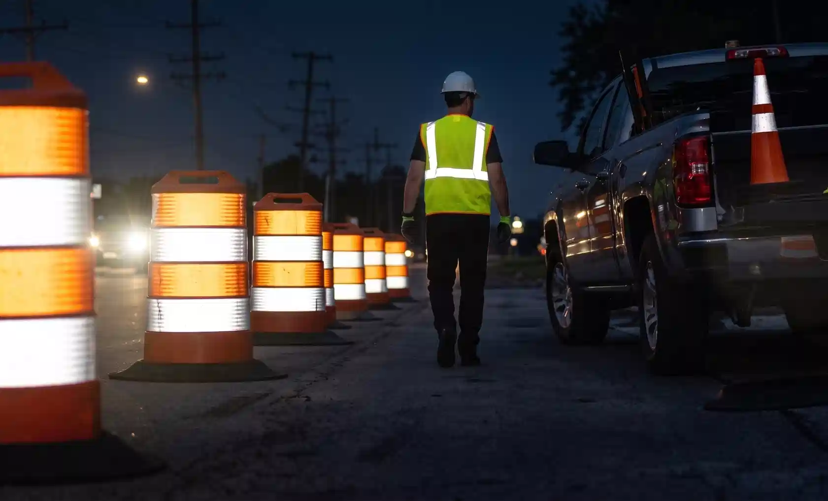 Nighttime construction scene showing a worker in high-vis vest and a line of intensely retro-reflective traffic drums.